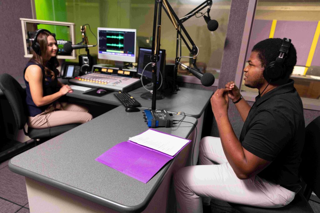Zacchaeus and a woman sit across from each other at a desk. On the desk is a computer and other various podcast equipment. They both speak into microphones.
