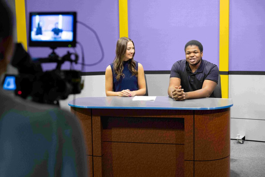 Zacchaeus and a woman sit at a table as a man records them with a professional camera. The woman smiles at Zacchaeus as he talks to the camera.