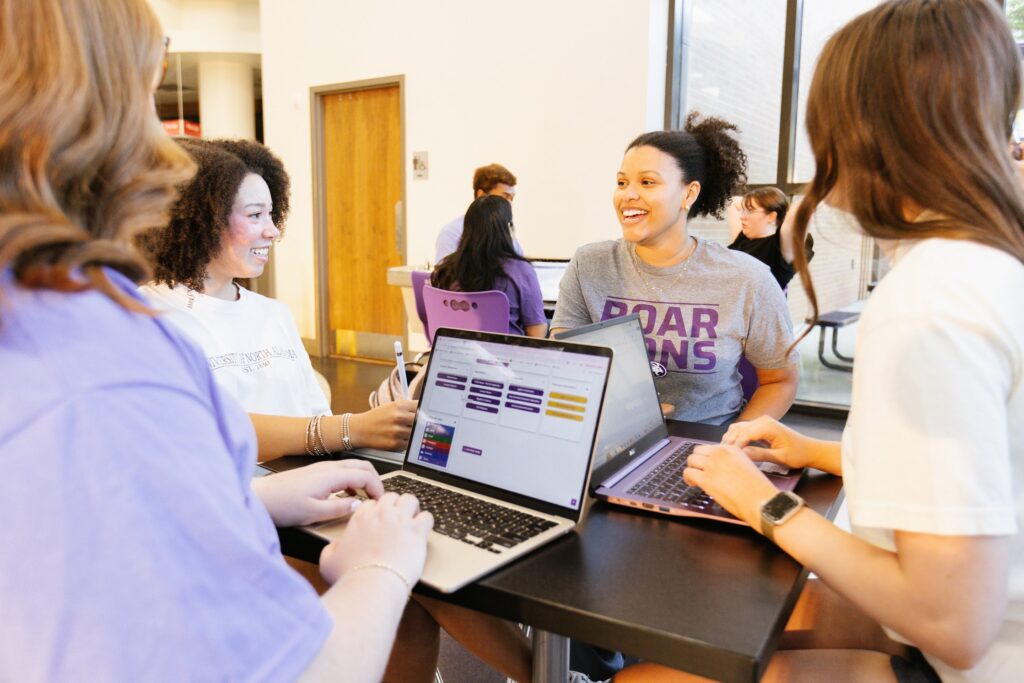 Krimsyn and three other students smile and laugh as they sit around a table at UNA. Two of them have laptops out.