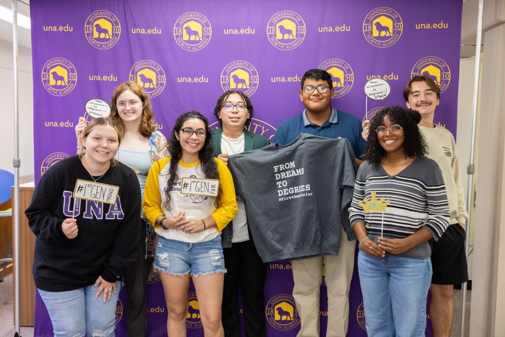 Micah smiles and poses against a UNA photo backdrop with six fellow first generation students.