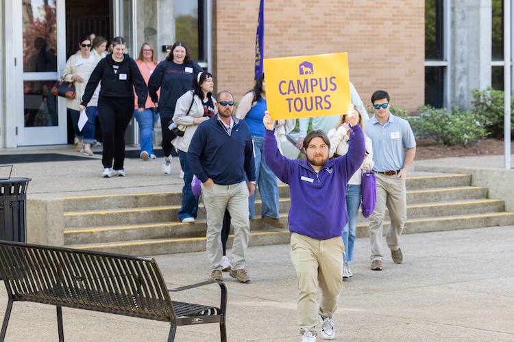 Oakley leads a group of people on UNA's campus. He holds up a sign with UNA's logo that reads "Campus Tours."