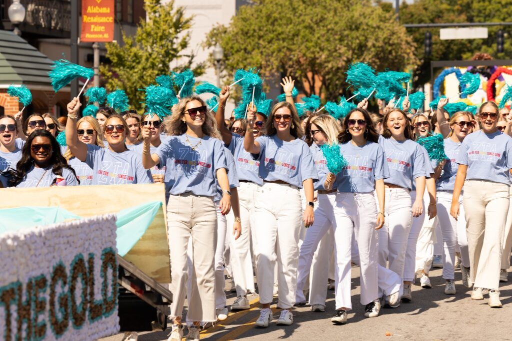 Sadye Webb and a large group of ZETA sorority members smile as they walk down the road, waving pompoms with long handles.