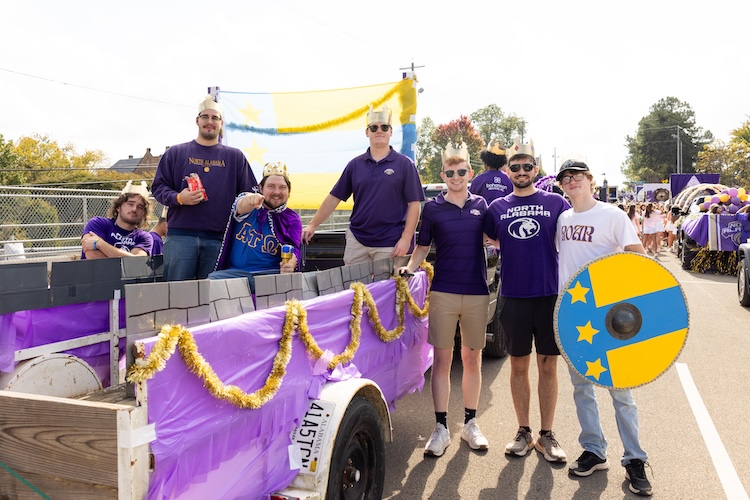 Oakley sits on a float while wearing a cape and crown. Six other students stand around him, both in and out of the float. Many of them are also wearing crowns.