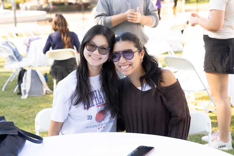 Sydney and another student sit at one of many tables outdoors for an event. They sit close and smile at the camera.